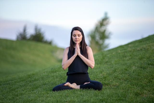A young woman sits cross-legged in a park. She is closing her eyes and appears to be meditating.
